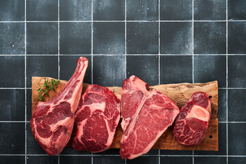 Wall Mural - Raw prime steaks. Variety of fresh black angus prime meat steaks T-bone, New York, Ribeye, Striploin, Tomahawk on black old tiles stone background. Set of various classic steaks. Top view copy space.