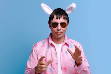 Foto de um homem positivo funky vestido de óculos de sol de fantasia de coelho rosa direcionando o espaço vazio mão na cintura isolada no fundo de cor azul
