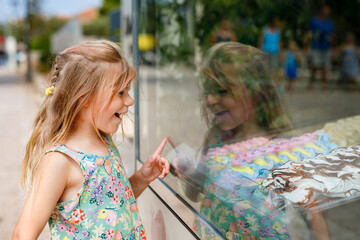 Wall Mural - Happy preschool girl choosing and buying ice cream in outdoor stand cafe. Cute child looking at different sorts of icecream. Sweet summer dessert on family vacations. Summertime.