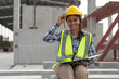 © Supachai - Asian woman engineer holding document smiling at construction site. Confident female Indian wearing protective helmet and vest setting in factory making precast concrete wall for real estate housing.