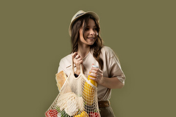 Wall Mural - Smiling young woman in light summer clothes with a mesh eco bag full of vegetables, greens watching in a camera on a green studio background. Sustainable lifestyle. Eco friendly concept. Zero waste.