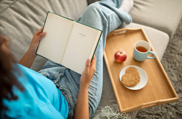 Millennial african american lady reading book, enjoy food and drink tea on sofa in living room