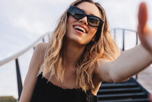 Cheerful Lady Making Selfie At Sunny Day. Pretty Young Girl With Curly Blonde Hair And Two Thin Braids Towards To Camera, Smiling And Wear Black Glasses And Dress Under The Stairs Bridge And Sky.