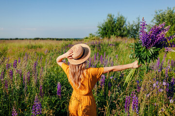 Wall Mural - Happy woman holding bouquet of lupin flowers walking in summer meadow. Girl opens arms enjoying landscape