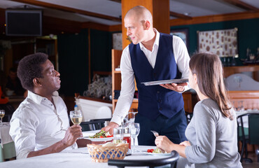 Wall Mural - Polite waiter serving adult biracial couple in restaurant, bringing ordered meals