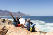 © Wavebreak Media - Happy senior biracial couple sitting on rocks by the sea, using smartphone and taking pictures