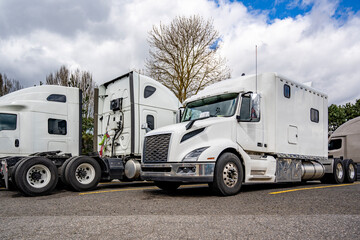 Three white big rigs semi trucks tractors without semi trailers and different cab lengths for a comfortable rest of the truck drivers stand side by side in the truck stop parking lot