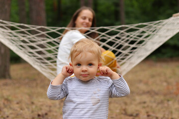 Wall Mural - Outdoor closeup shot of cute blonde infant baby playing with mother sitting in hammock, family relaxing in forest, plaing in open air, enjoying summer days.