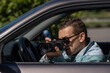 © Volha - Hidden photographing. Paparazzi Concep Photographer uses his professional camera. Private detective man in car window. Mobile reporter. Photojournalist at work. Detective or investigator.