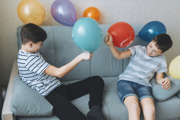 Happy teenager boys playing and having fun with balloons sitting on couch at home. Two smiling joyful playful friends enjoying entertaining fooling around on birthday party indoors. Carefree childhood