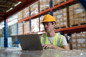 Male warehouse worker wearing uniform checks stock inventory in warehouse. Male worker using laptop computer and checking barcodes on boxes on shelf pallet in storage warehouse