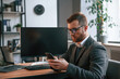 © standret - Sitting, using smartphone. Businessman in suit is in the office with computer