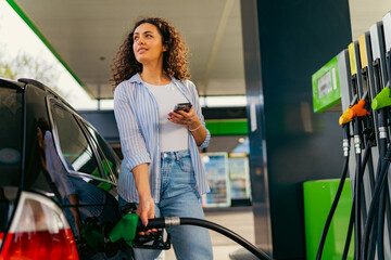 A young beautiful woman does not follow the safety rules and uses the phone while filling her car gas tank