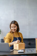 © ArLawKa - Asian businesswoman using smartphone to start online delivery Freelancer or salesman checking production order with laptop. SME entrepreneur working with parcel box at home.