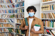 © Liubomir - Portrait of a young male student inside a library studying at a university, hispanic man with curly hair looking at the camera holding books in his hands, the guy is wearing a protective face mask.