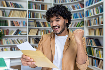 Young student received letter with good exam results and university admission confirmation, hispanic man with curly hair celebrating successful achievement holding hand up close up.