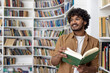 © Liubomir - Young hispanic student reading a book while standing inside an academic university library, man smiling contentedly looking towards the window.