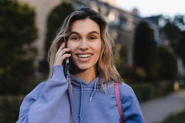 Wall Mural - Happy blonde curly woman talking phone and look happy, smiling. Portrait of gorgeous smiling female using mobile phone. Girl wear purple hoody and pink bag.