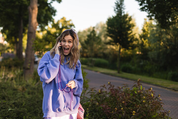 Wall Mural - Happy blonde curly woman talking phone and look happy, smiling. Portrait of gorgeous smiling female using mobile phone. Girl wear purple hoody and pink bag, look at side, surprised and shocked.