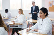 © JackF - Young positive woman in white medical coat sitting at desk in classroom attending seminar