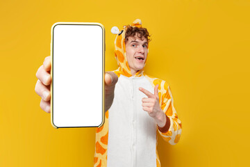 young curly guy in funny giraffe pajamas shows blank smartphone screen on yellow background