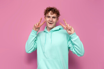 young curly guy in mint hoodie shows gesture of peace with his hands on pink isolated background