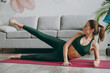 © ViDi Studio - Full body side view young strong sporty athletic fitness trainer instructor woman wear green tracksuit lying on side raises legs train do exercises at home gym indoor Workout sport motivation concept.