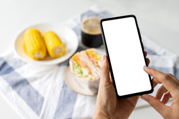 Woman enjoying healthy breakfast at home, hand holding a smartphone with blank screen for mockup design