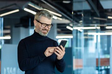 Mature adult man inside office standing near window, businessman typing message on phone and browsing internet pages, gray-haired boss smiling reading news online.