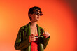 trendy woman in green leather jacket posing in several sunglasses and looking away on orange background.