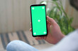 Young man sitting at home holding smartphone green mock-up screen in hand. Male person using chroma key mobile phone. Vertical mode. Touching, swiping display, tapping, surfing Internet social media