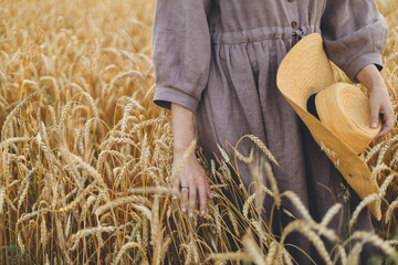 Woman with straw hat holding wheat stems in field. Atmospheric tranquil moment. Female in rustic linen dress touching ripe wheat ears in evening summer countryside. Rural slow life