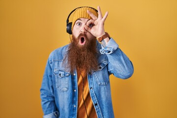 Wall Mural - Caucasian man with long beard listening to music using headphones doing ok gesture shocked with surprised face, eye looking through fingers. unbelieving expression.