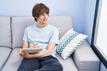 Sticker - Young blond man writing on notebook sitting on sofa at home