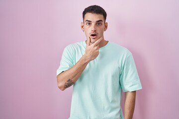 Wall Mural - Handsome hispanic man standing over pink background looking fascinated with disbelief, surprise and amazed expression with hands on chin