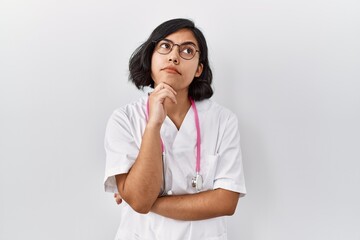 Wall Mural - Young hispanic doctor woman wearing stethoscope over isolated background with hand on chin thinking about question, pensive expression. smiling with thoughtful face. doubt concept.