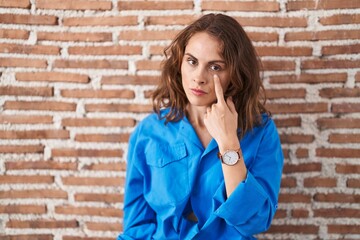 Poster - Beautiful brunette woman standing over bricks wall pointing to the eye watching you gesture, suspicious expression