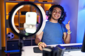 Canvas Print - Hispanic man with curly hair playing video games recording with smartphone smiling happy and positive, thumb up doing excellent and approval sign