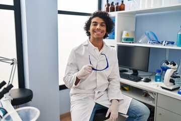 Poster - Young hispanic man wearing scientist uniform holding safety glasses at laboratory