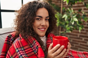 Wall Mural - Young beautiful hispanic woman drinking coffee covering with blanket at home