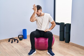 Sticker - Hispanic man with beard sitting on pilate balls at yoga room surprised with hand on head for mistake, remember error. forgot, bad memory concept.