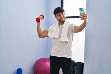 Canvas Print - Young hispanic man holding dumbbell make selfie by smartphone at sport center