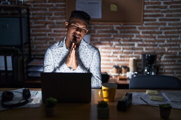 Wall Mural - Young hispanic man working at the office at night begging and praying with hands together with hope expression on face very emotional and worried. begging.