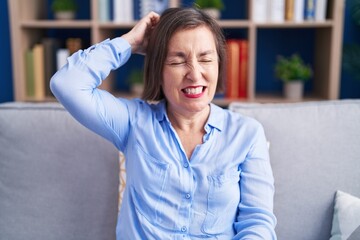 Sticker - Middle age hispanic woman sitting on the sofa at home confuse and wonder about question. uncertain with doubt, thinking with hand on head. pensive concept.