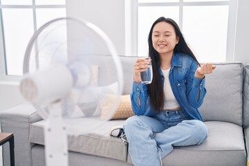 Poster - Young asian woman drinking glass of water enjoying air from fan screaming proud, celebrating victory and success very excited with raised arm