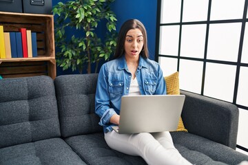 Wall Mural - Hispanic woman using laptop at home scared and amazed with open mouth for surprise, disbelief face