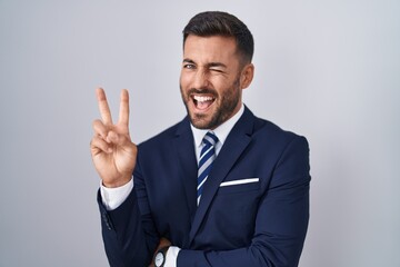 Wall Mural - Handsome hispanic man wearing suit and tie smiling with happy face winking at the camera doing victory sign with fingers. number two.