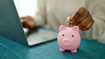 Canvas Print - African american woman using laptop inserting coin on piggy bank at dinning room