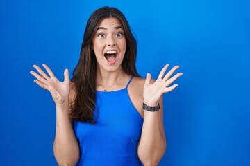 Poster - Hispanic woman standing over blue background celebrating crazy and amazed for success with arms raised and open eyes screaming excited. winner concept