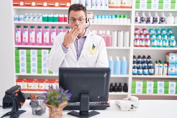 Poster - Young hispanic man working at pharmacy drugstore wearing headset covering mouth with hand, shocked and afraid for mistake. surprised expression